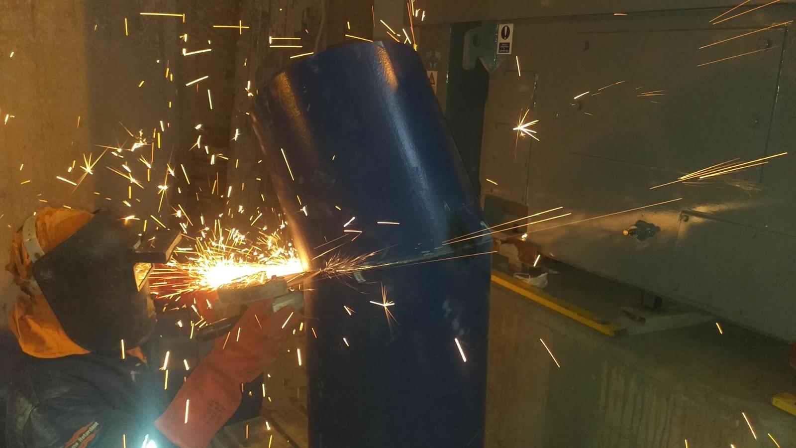 Technician grinding a blue fabricated pipe fitting with sparks across the workshop