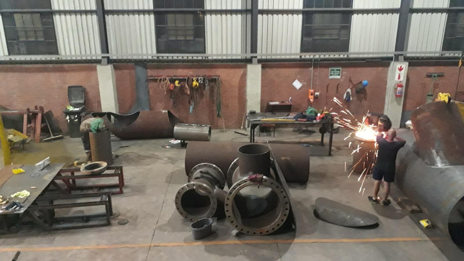 Wide workshop view with large steel pipe fittings and a worker grinding a component