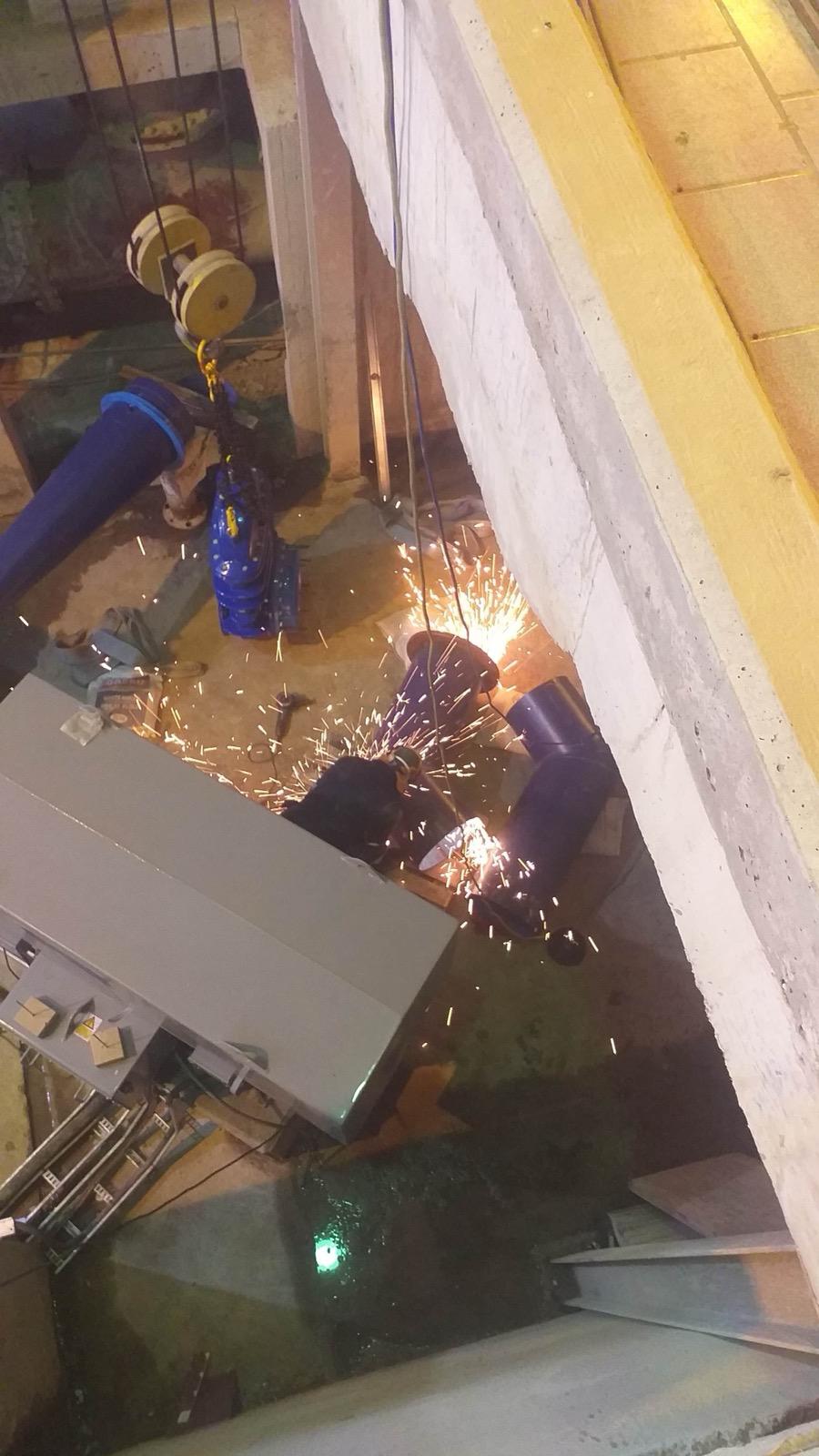 Technicians cutting or grinding blue flanged pipework beside a valve and lifting equipment